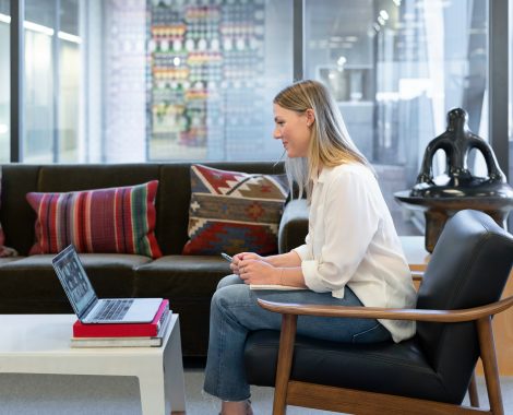 Woman sitting in a modern office space, smiling and looking at a laptop screen during a video call or virtual meeting.