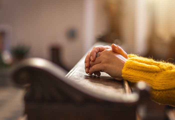 Close-up of hands clasped in prayer resting on a wooden pew, with a person wearing a yellow sweater in a quiet setting.