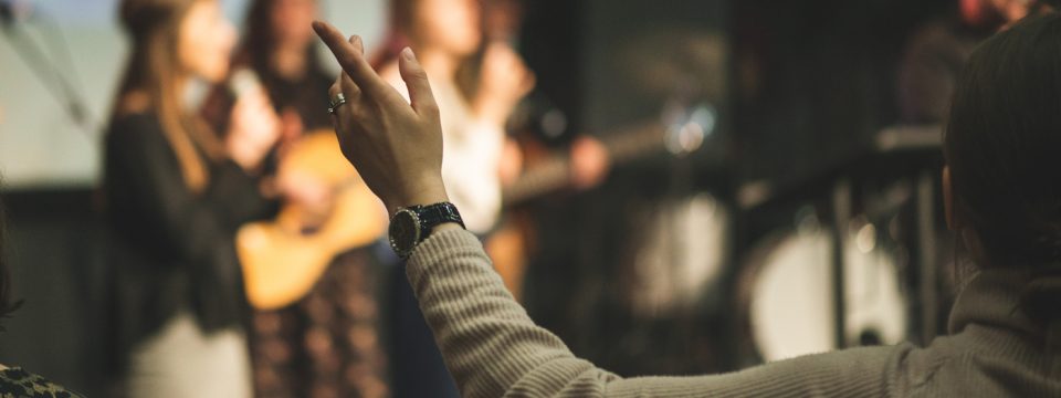 Person raising a hand in worship during a live music performance or religious service, with a band playing in the background.