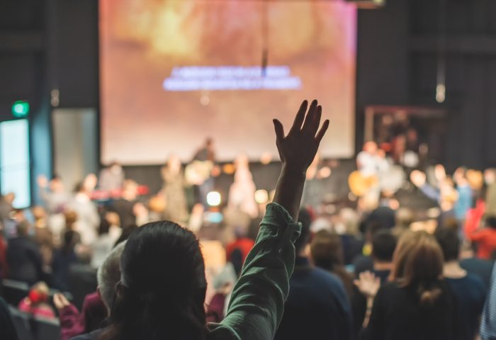 Person raising their hand in a large gathering or event, with a stage and audience in the background.